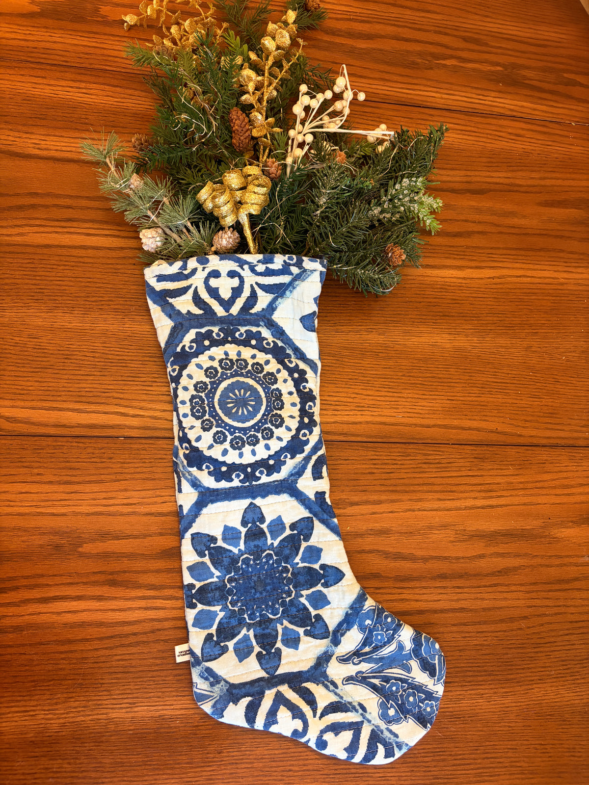 A blue and white patterned Christmas stocking filled with dried greenery and pinecones, laid flat on a warm brown wooden surface.
