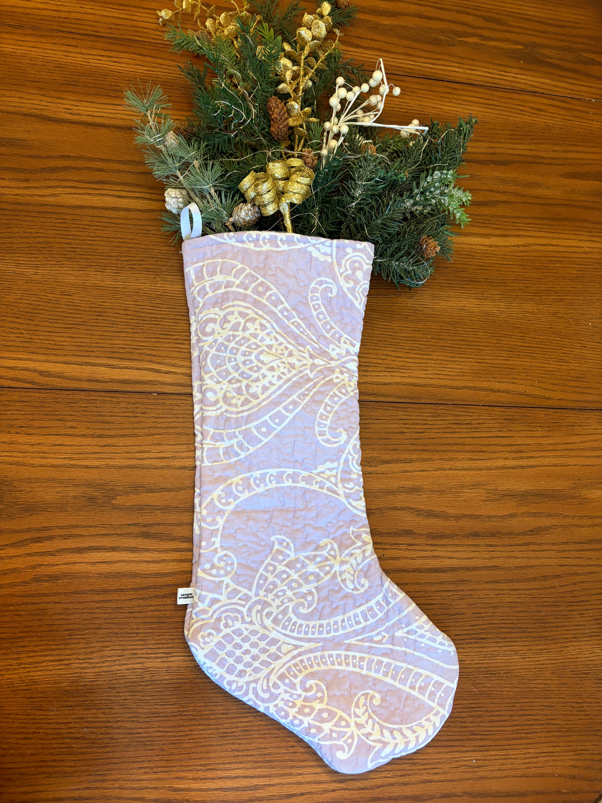 A light lavender Christmas stocking with a cream paisley pattern, filled with green pine branches, gold pinecones, and white berries, resting on a brown wooden surface.
