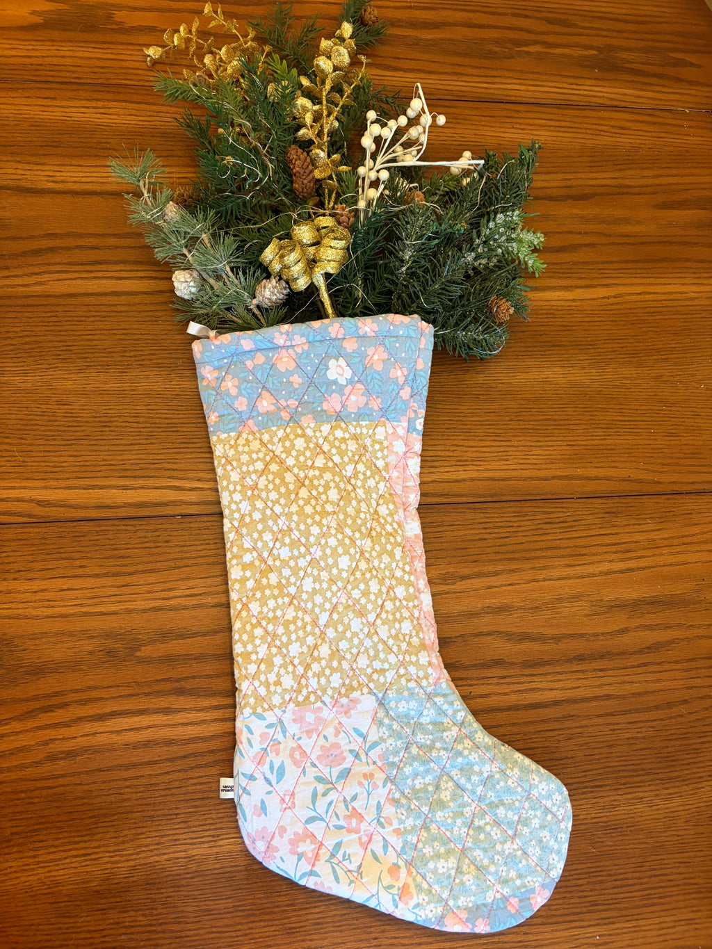 A handmade quilted Christmas stocking with a patchwork floral pattern in pastel blue, yellow, and pink, filled with green pine branches and dried flowers, laid flat on a wooden surface.
