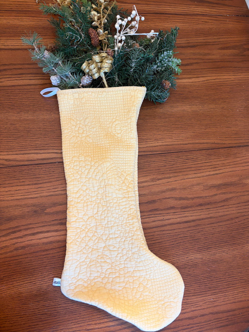A pale yellow quilted Christmas stocking with a textured pattern, filled with green pine branches, gold pinecones, and white berries, laid flat on a brown wooden surface.